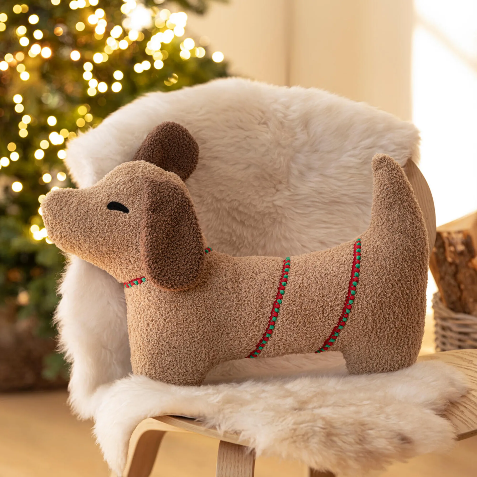 Dekokissen in Form eines Dackels auf einem Sessel mit weißem Kunstfell, vor einem beleuchteten Weihnachtsbaum im Hintergrund