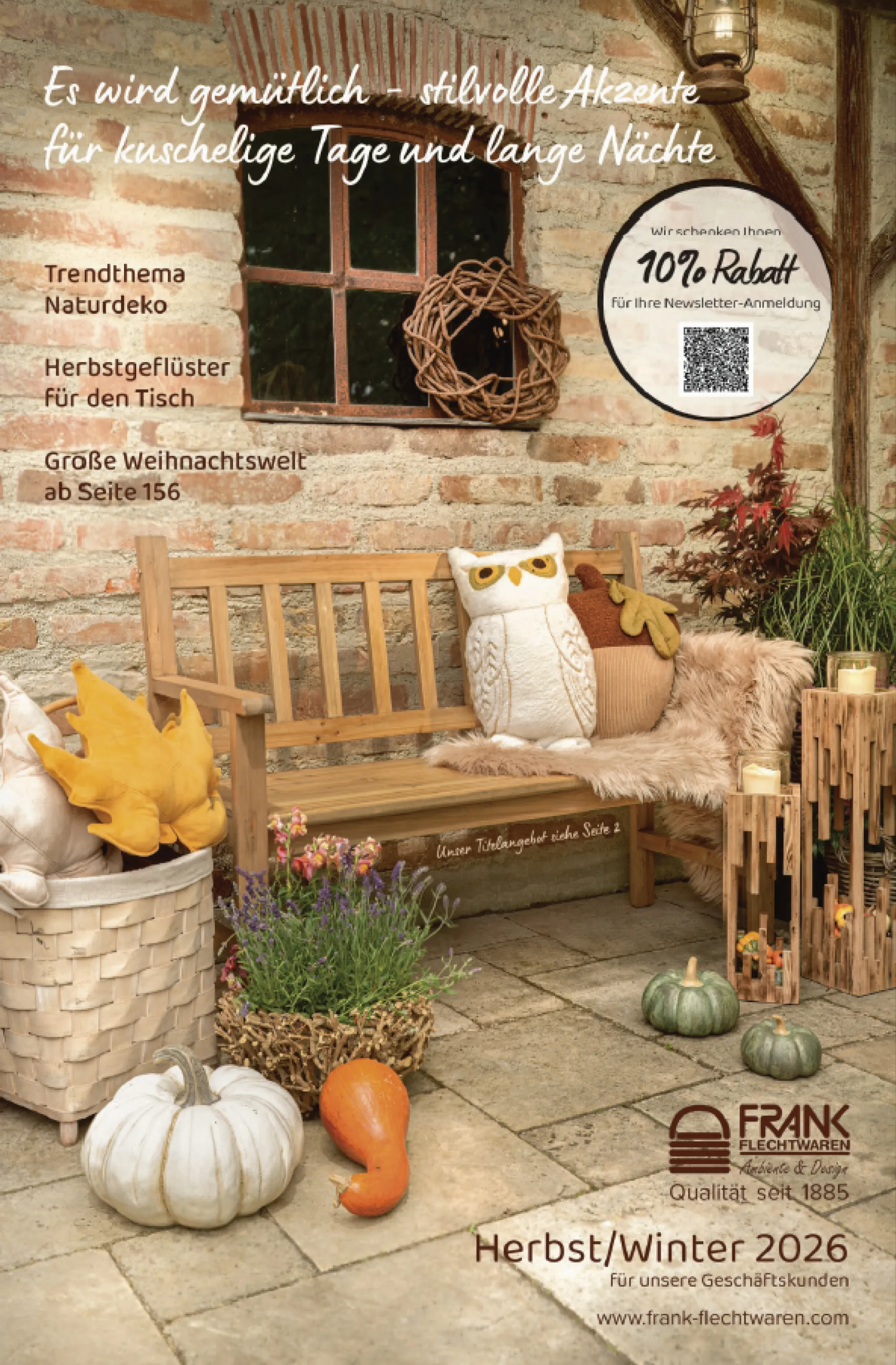 Herbstliche Outdoor-Dekoration mit Holzbank, Kürbissen, Eulenkissen, Holzwindlichtern, Flechtkorb und Pflanzen vor einer rustikalen Backsteinwand, Titelmotiv Katalog Herbst/Winter 2026 für Geschäftskunden